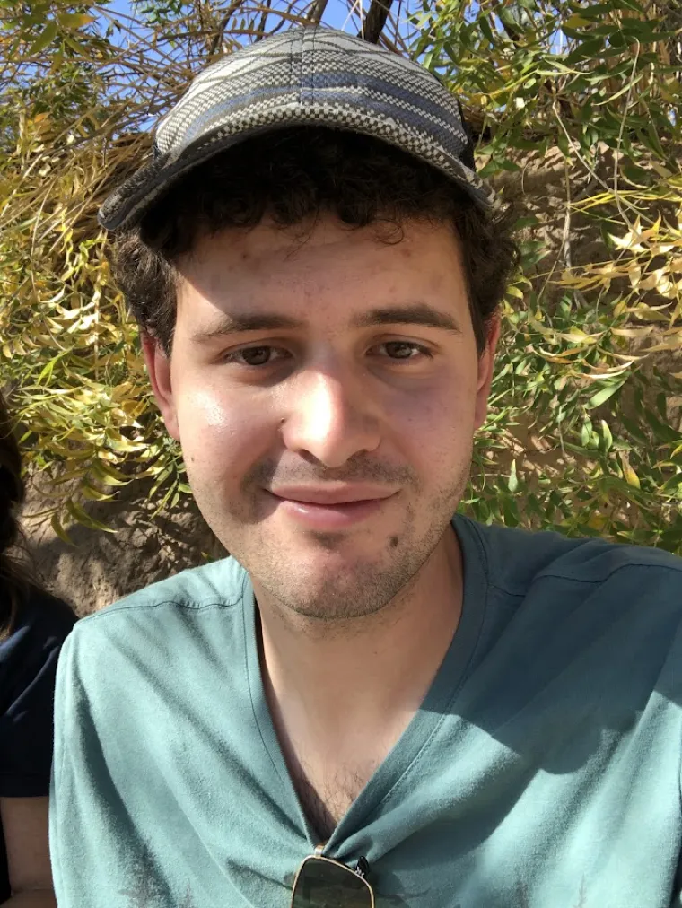 Myles Kashanipour: Man with dark curly hear wearing a grey hat and teal shirt. Background is a bush or a tree close up.